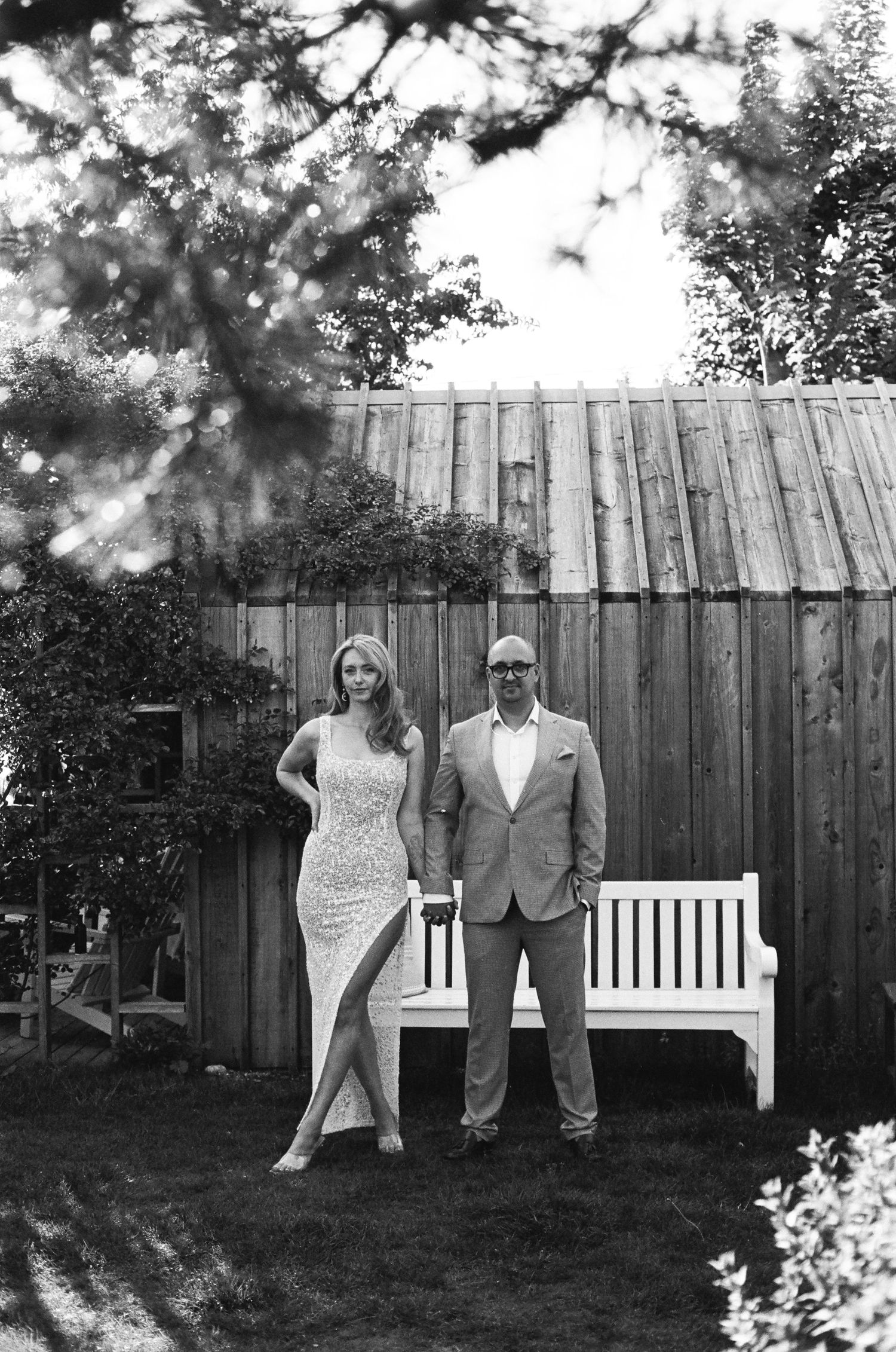 A couple poses together in four similar black and white portraits against a wooden fence in a garden setting.
