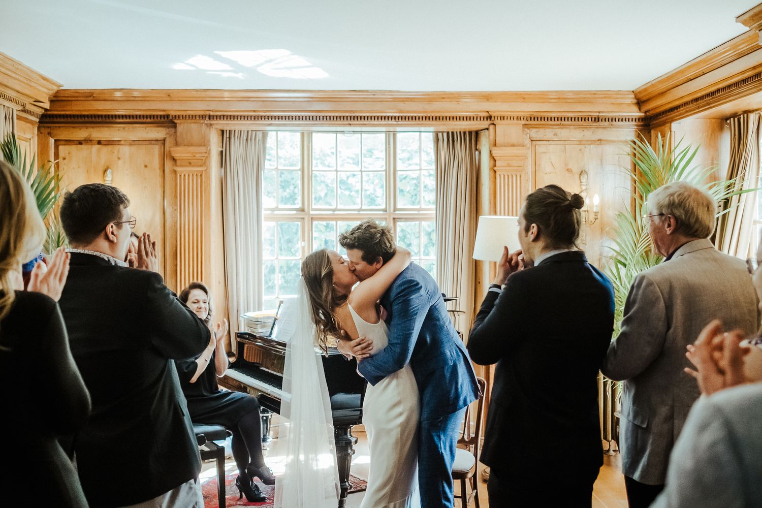 Wedding guests watch as newlyweds share their first kiss in an elegant wood-paneled room.