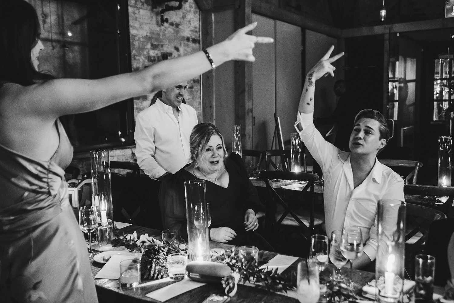 Black and white candid shot of people gesturing and celebrating at a dinner table with candles.