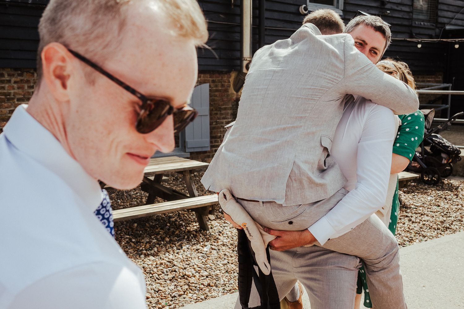 Wedding guests share an intimate embrace while another person in sunglasses stands nearby.