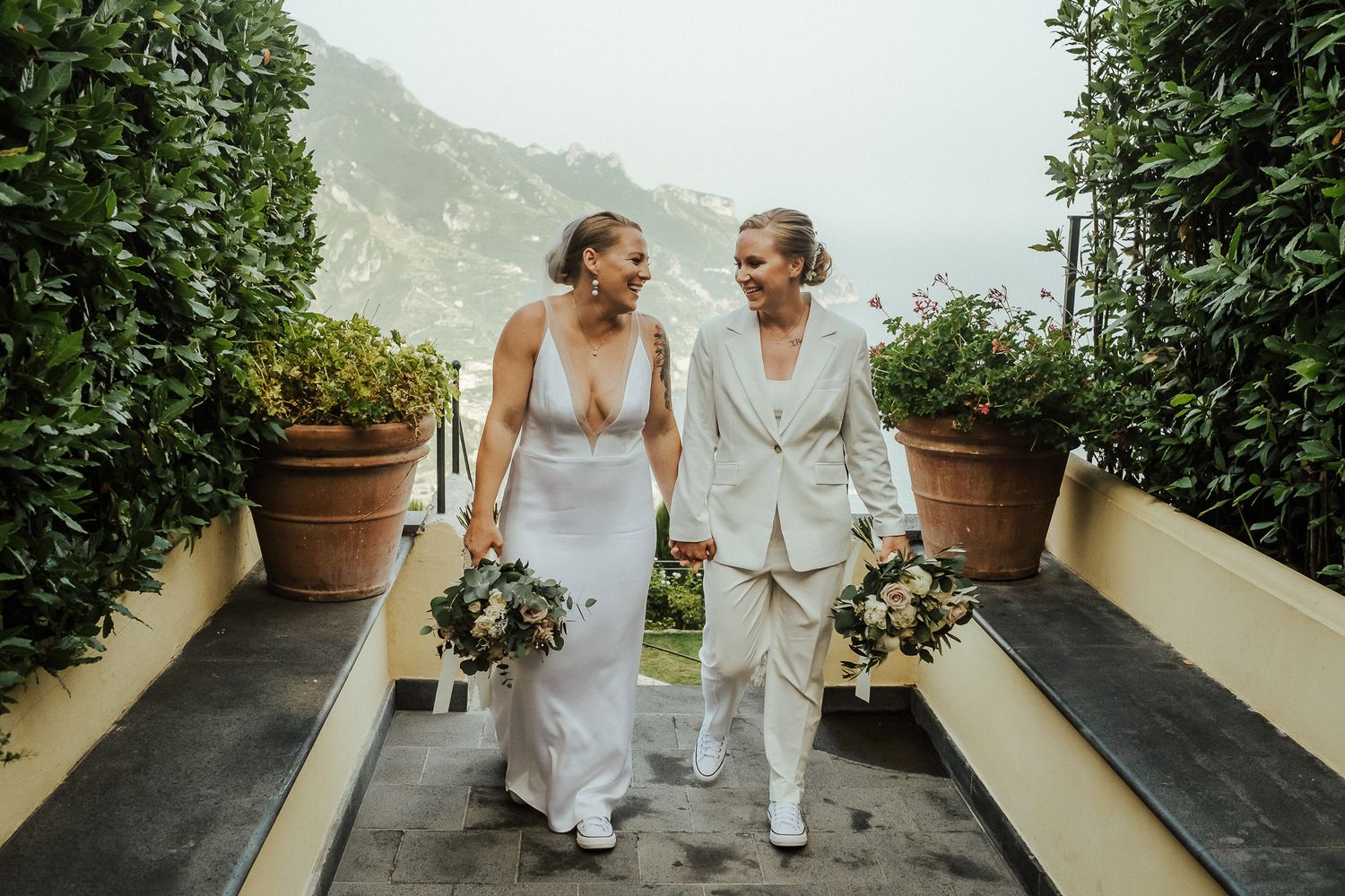 Two wedding couples walk hand in hand on a stone path surrounded by potted plants with mountains in background.