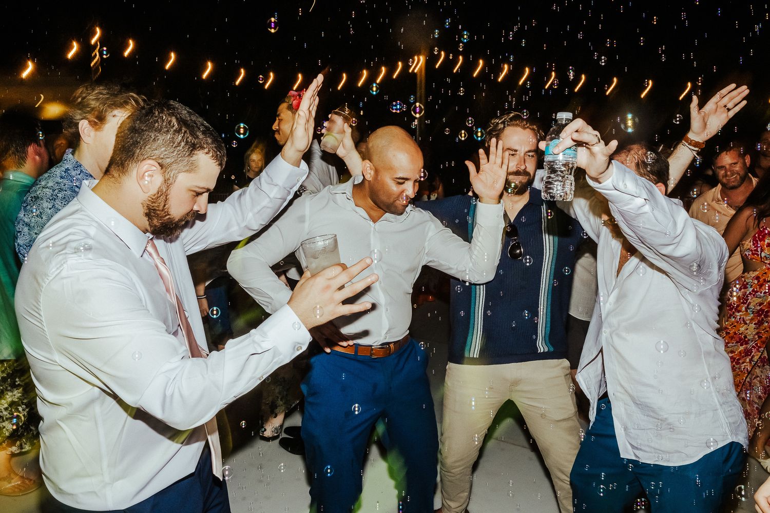 Group of partygoers dancing energetically with their arms raised under string lights at night.