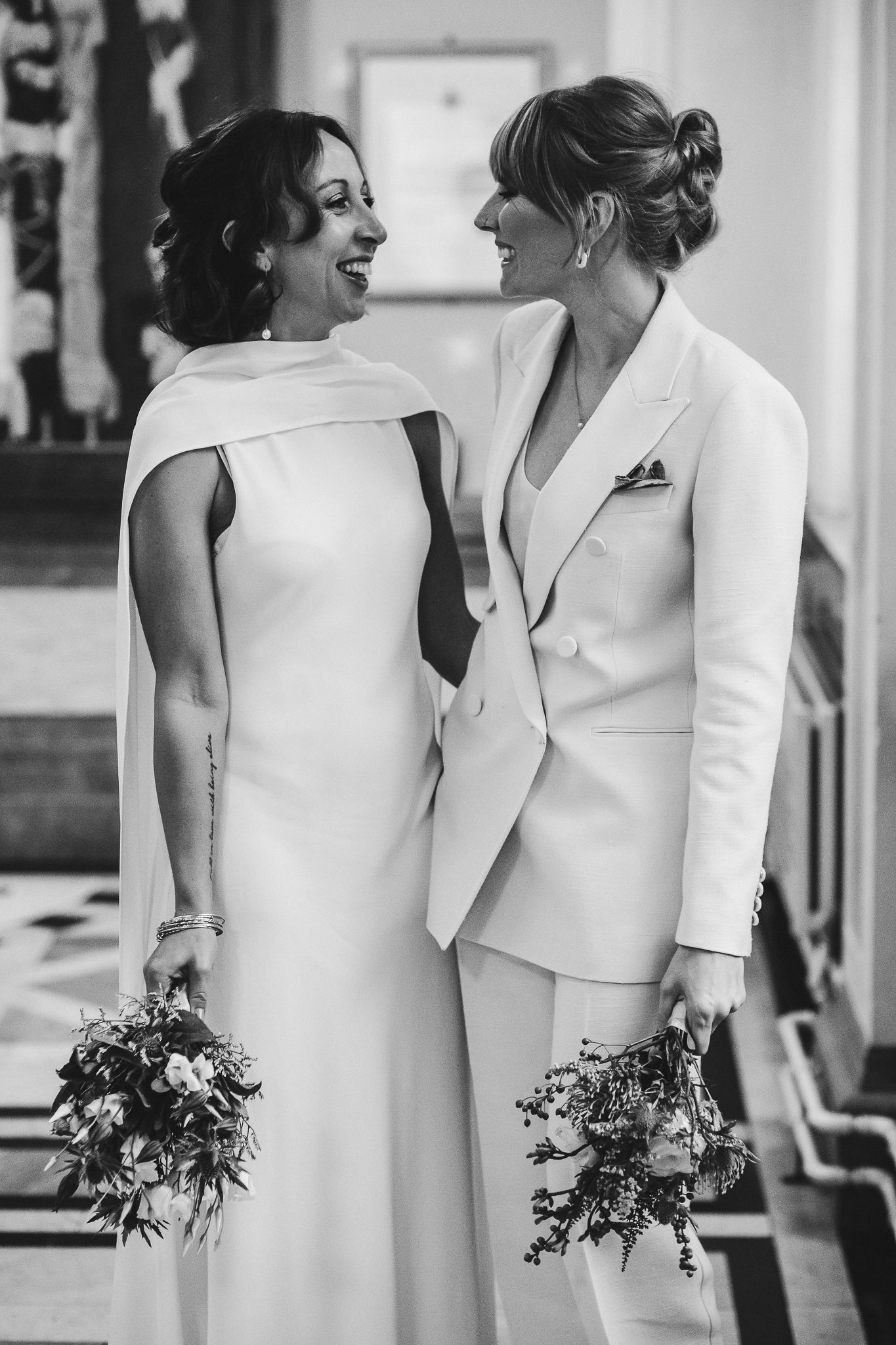 Two individuals in stylish white outfits share a joyful moment while holding bouquets.