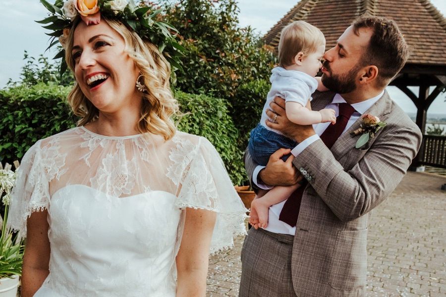 Joyful wedding moment with bride laughing while groom kisses baby.