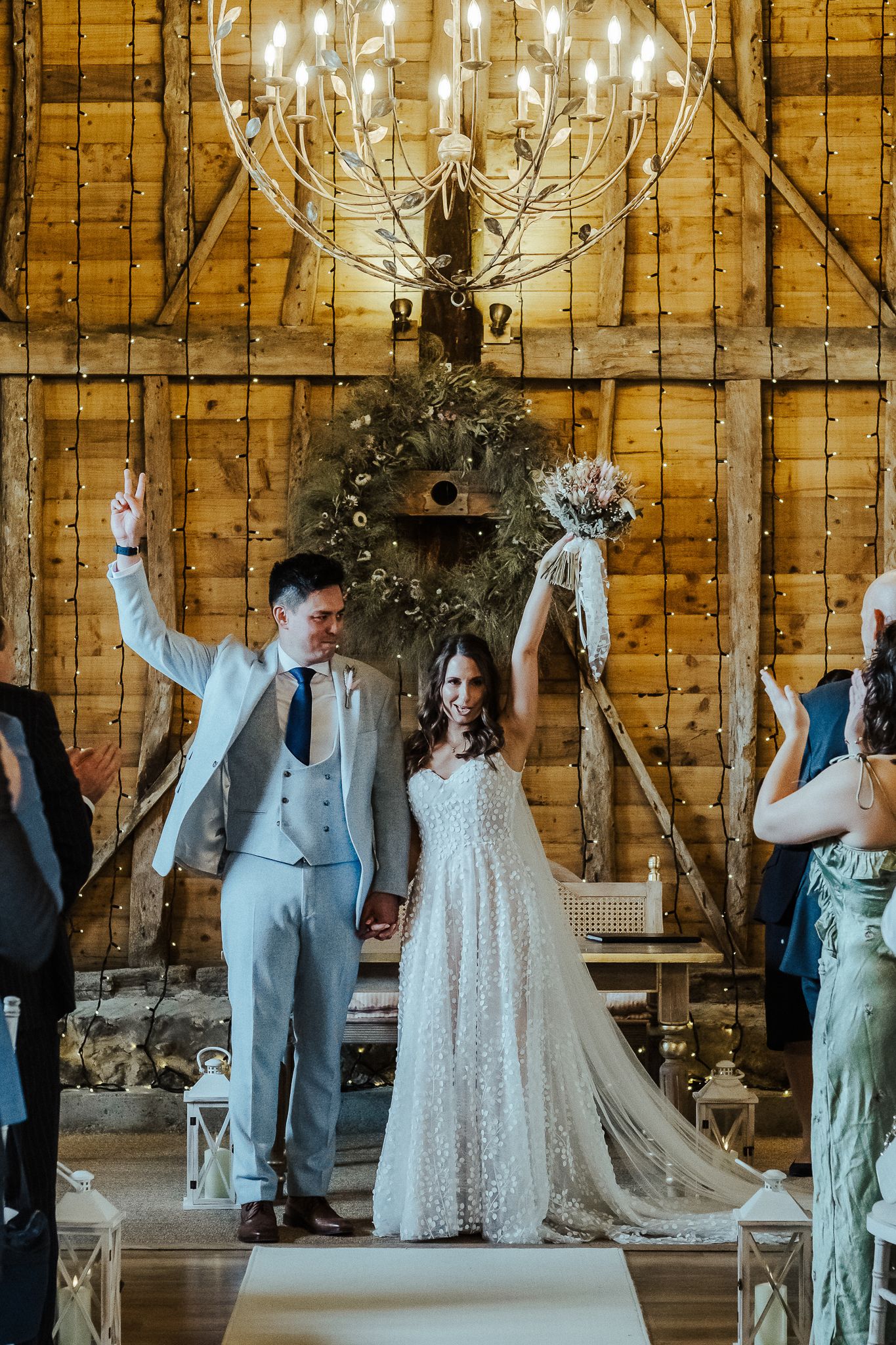 Newlyweds celebrate with raised arms under ornate chandelier in rustic barn venue.