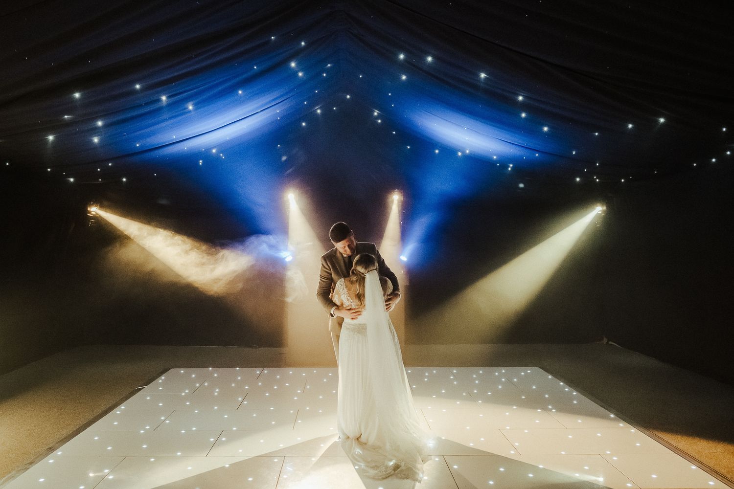 Couple shares first dance under dramatic stage lighting on illuminated dance floor.