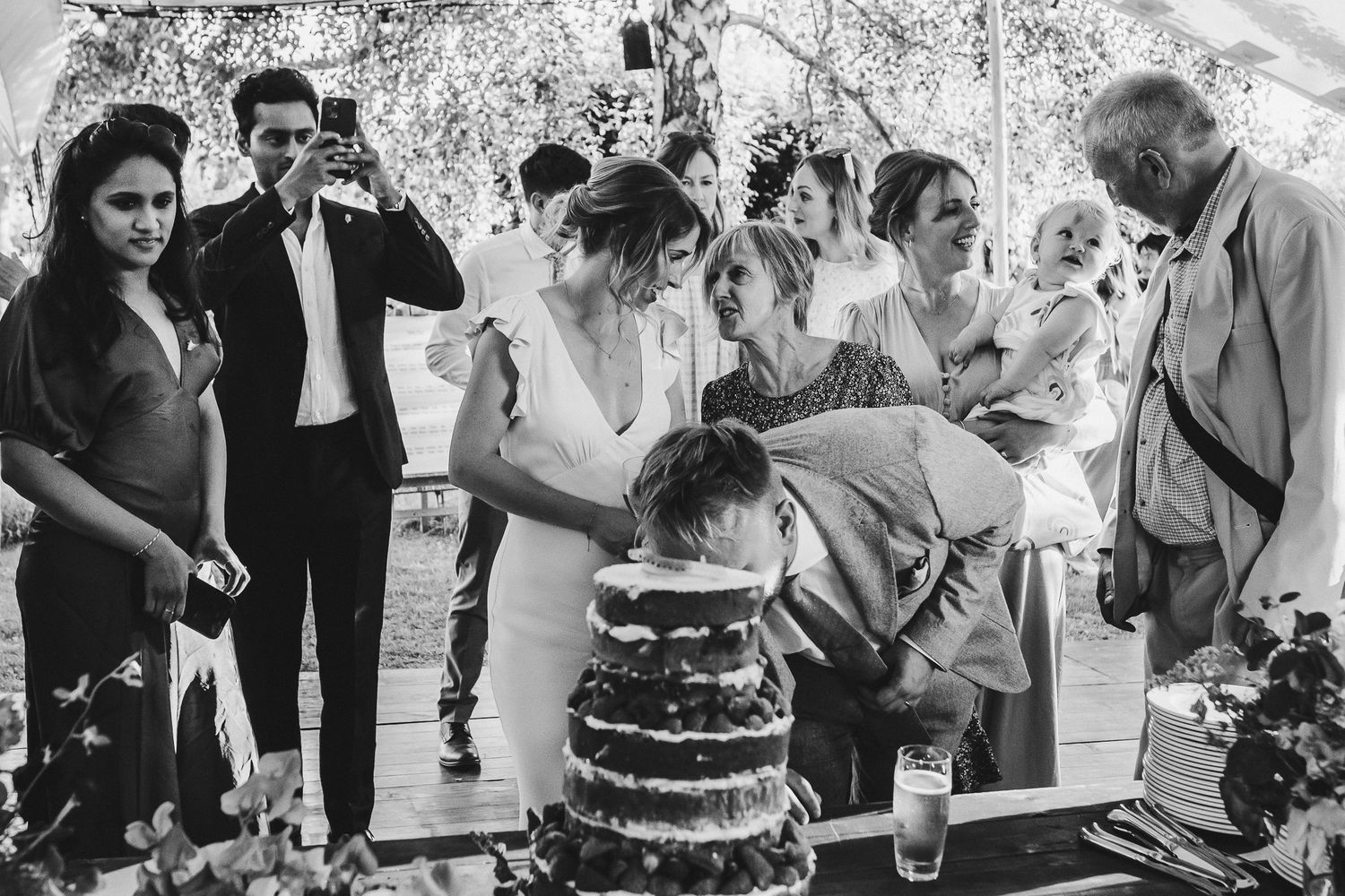Wedding guests gather around a rustic naked cake during outdoor reception.