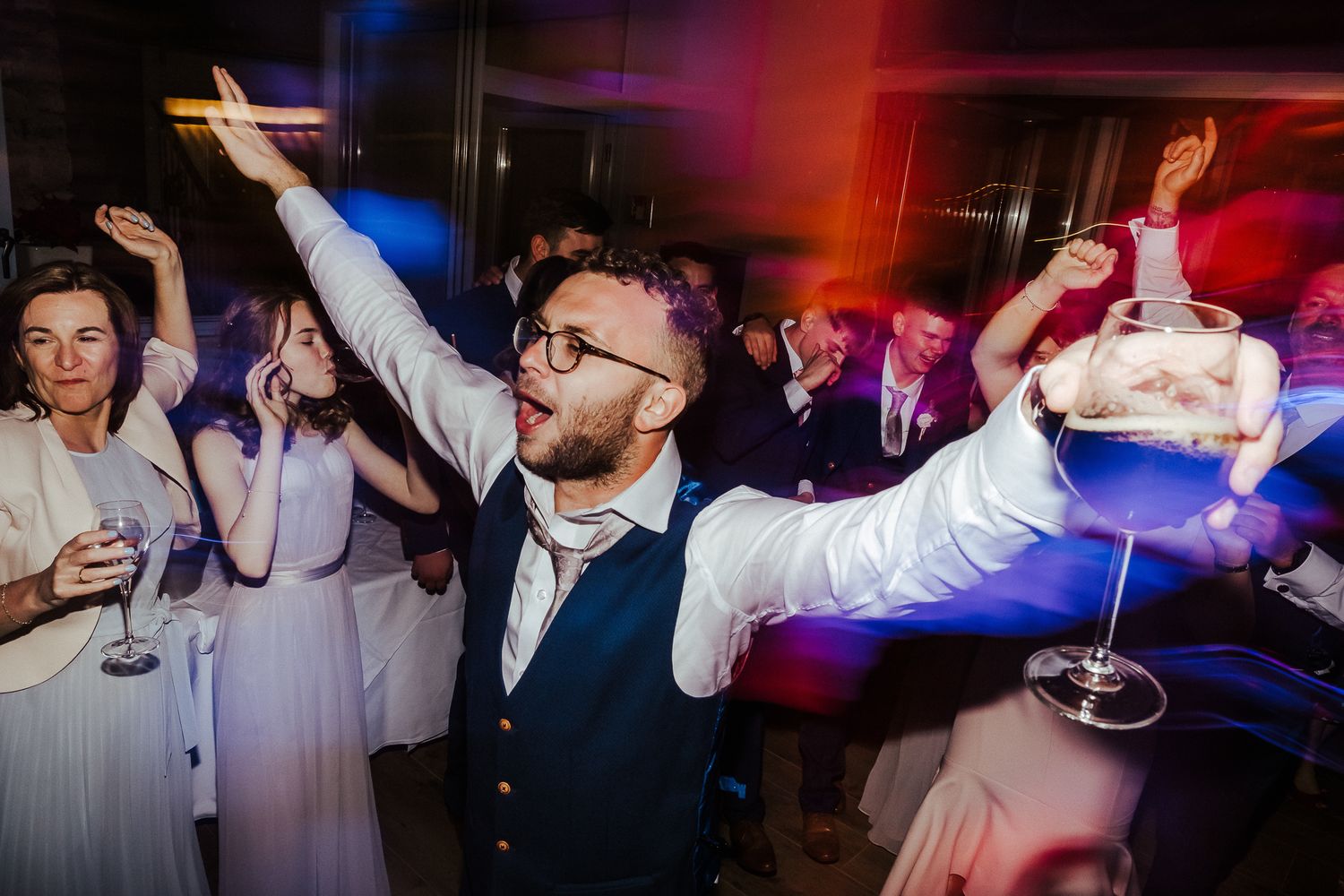 Energetic party guests dancing and celebrating with drinks under colorful reception lighting.
