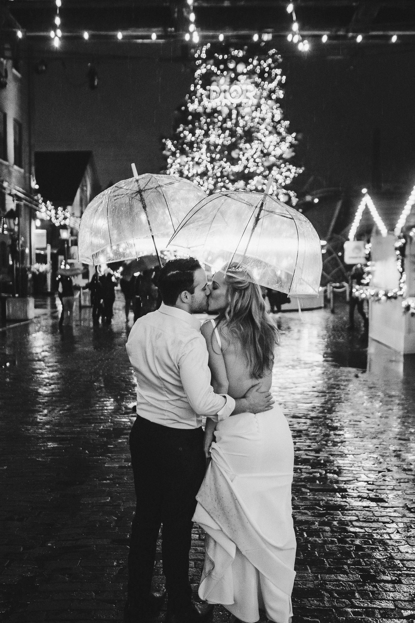 Couple shares romantic moment under clear umbrella with Christmas lights in background.