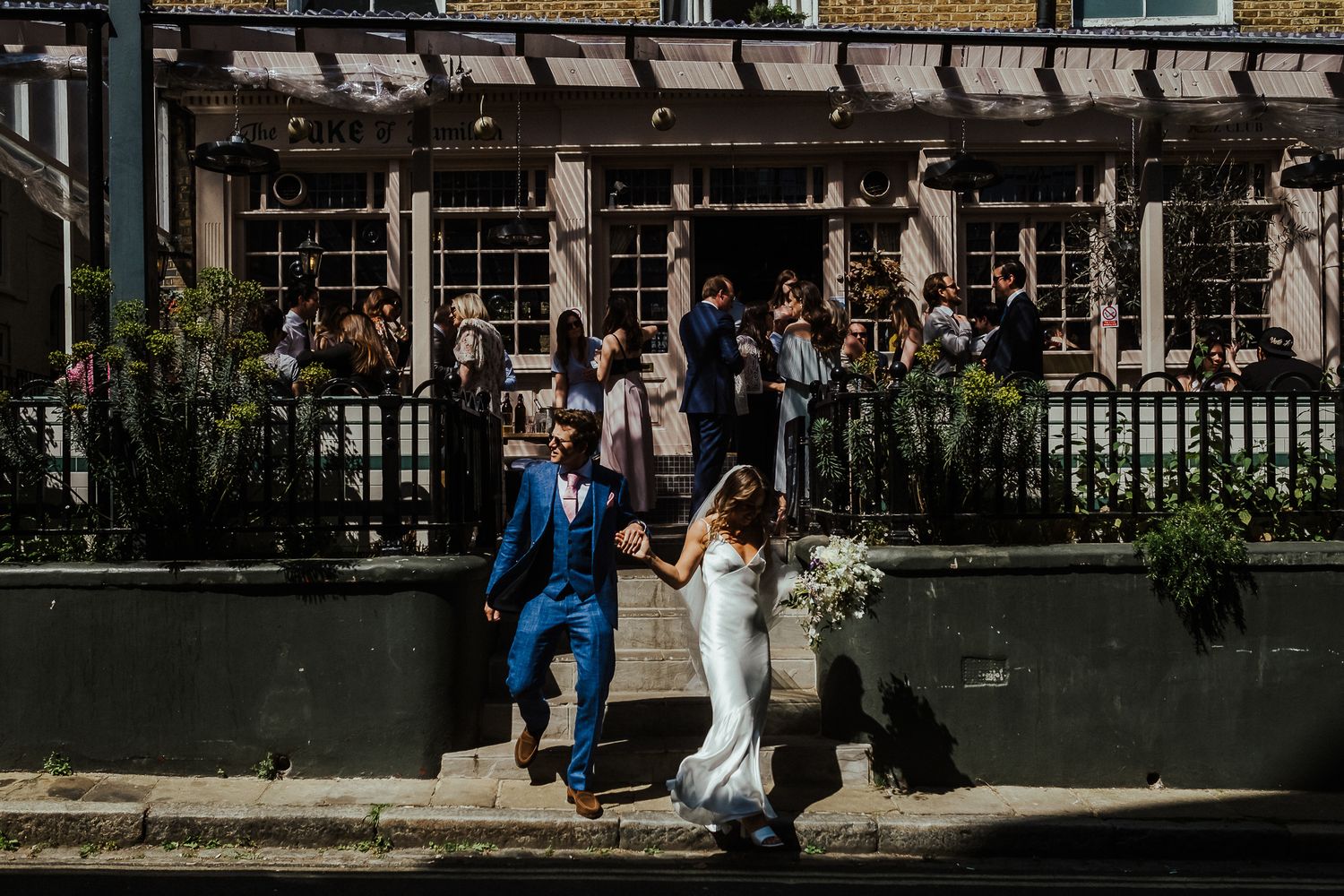 A wedding couple jumps joyfully outside a historic building with a blue suit and white dress.