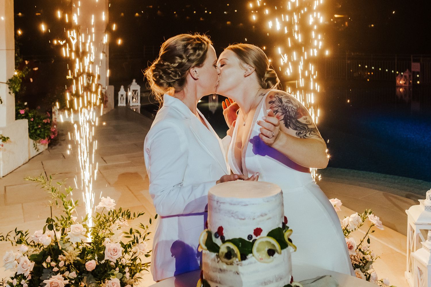 Couple shares intimate moment while cutting their wedding cake with dramatic sparkler fountains in background.