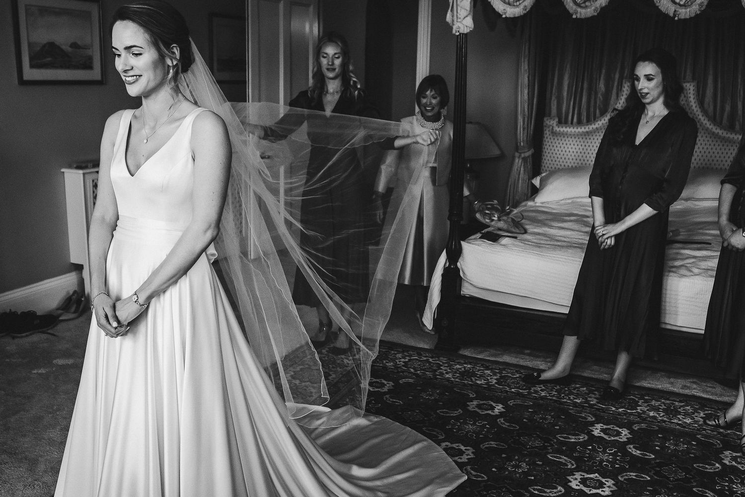 Bride in flowing white gown and veil stands while attendants look on.