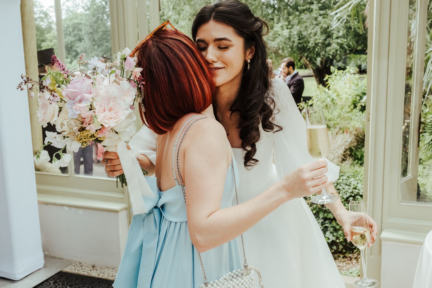 Wedding party mingling outdoors with champagne glasses in a garden setting.