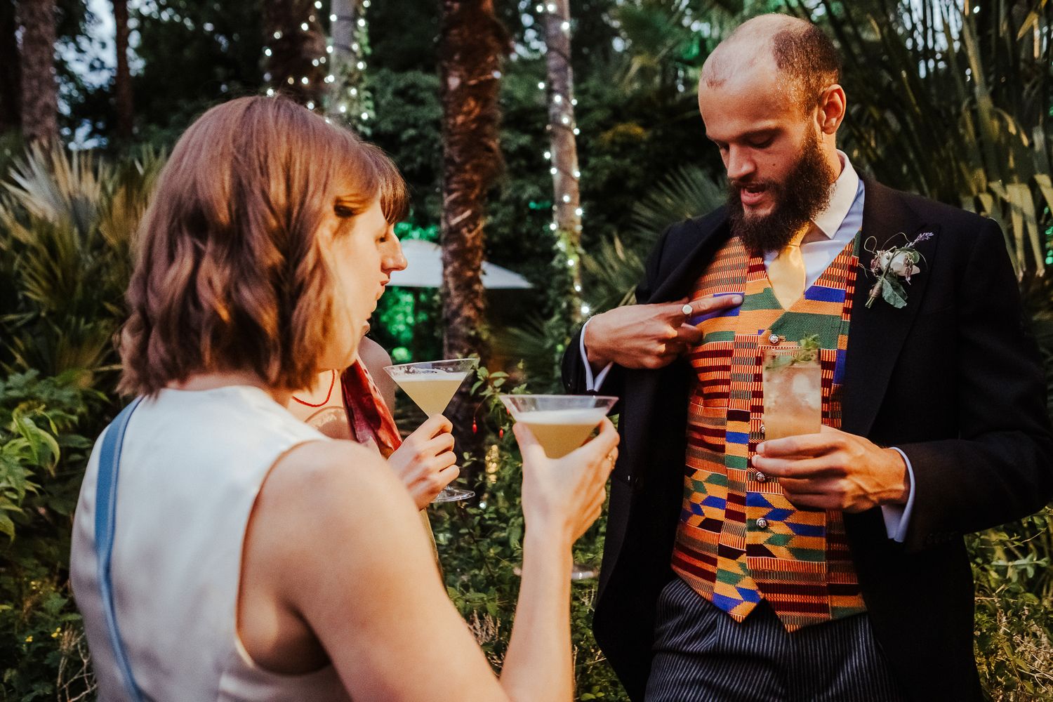 Two people share drinks and laughter in an outdoor garden setting during a festive celebration.