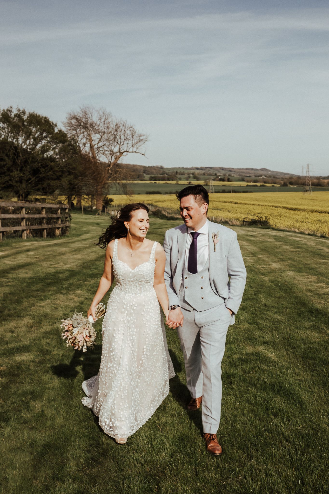 Couple walks hand in hand through countryside field in wedding attire.