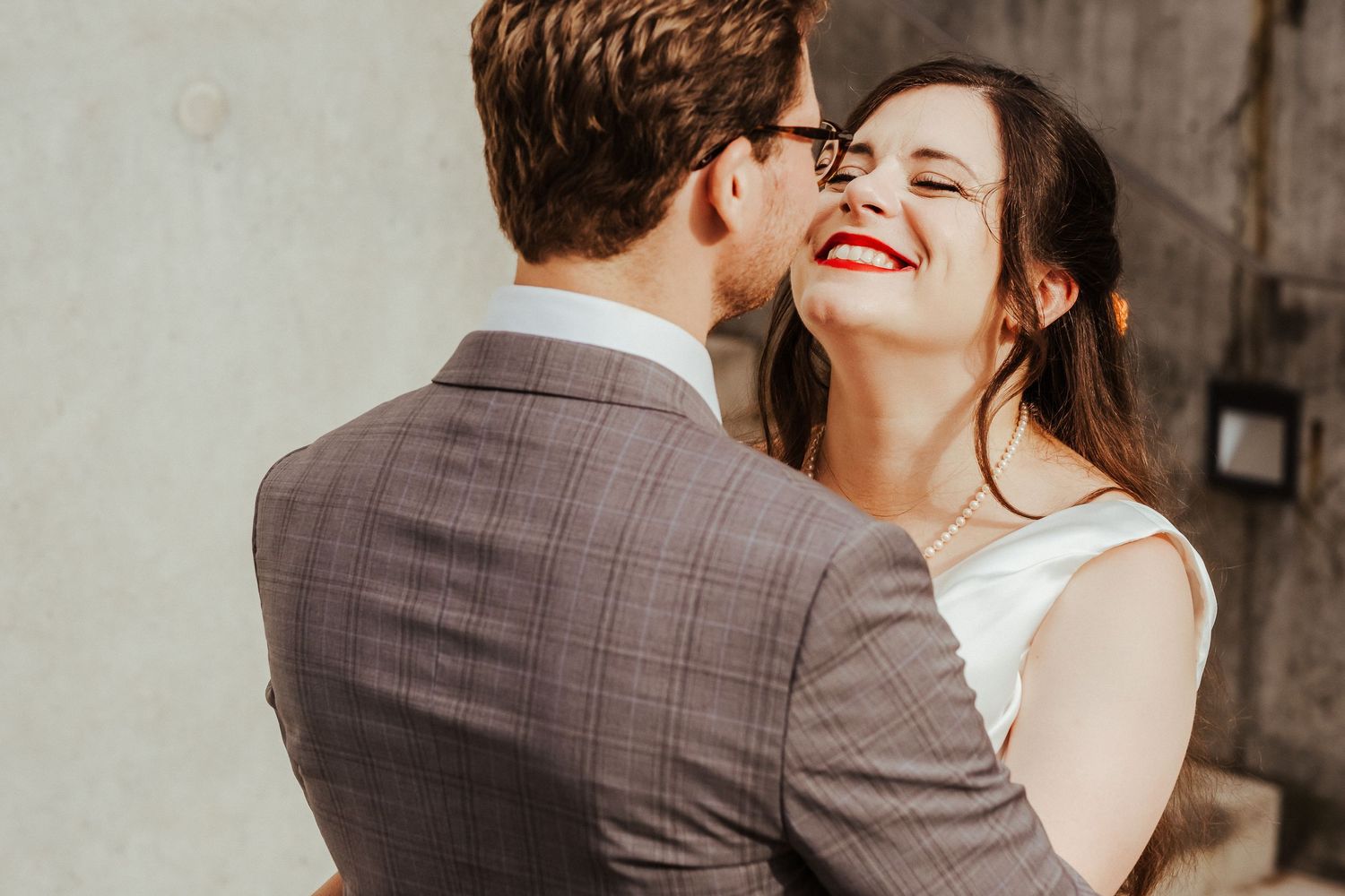 Intimate candid moment between newlyweds laughing against white wall.