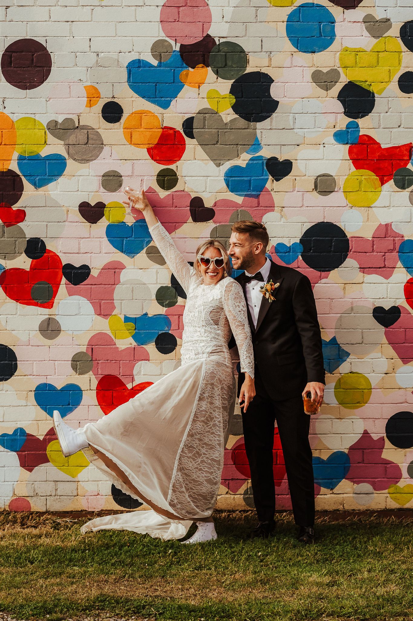Wedding couple posing against colorful heart mural with bride in lace dress and groom in tuxedo.