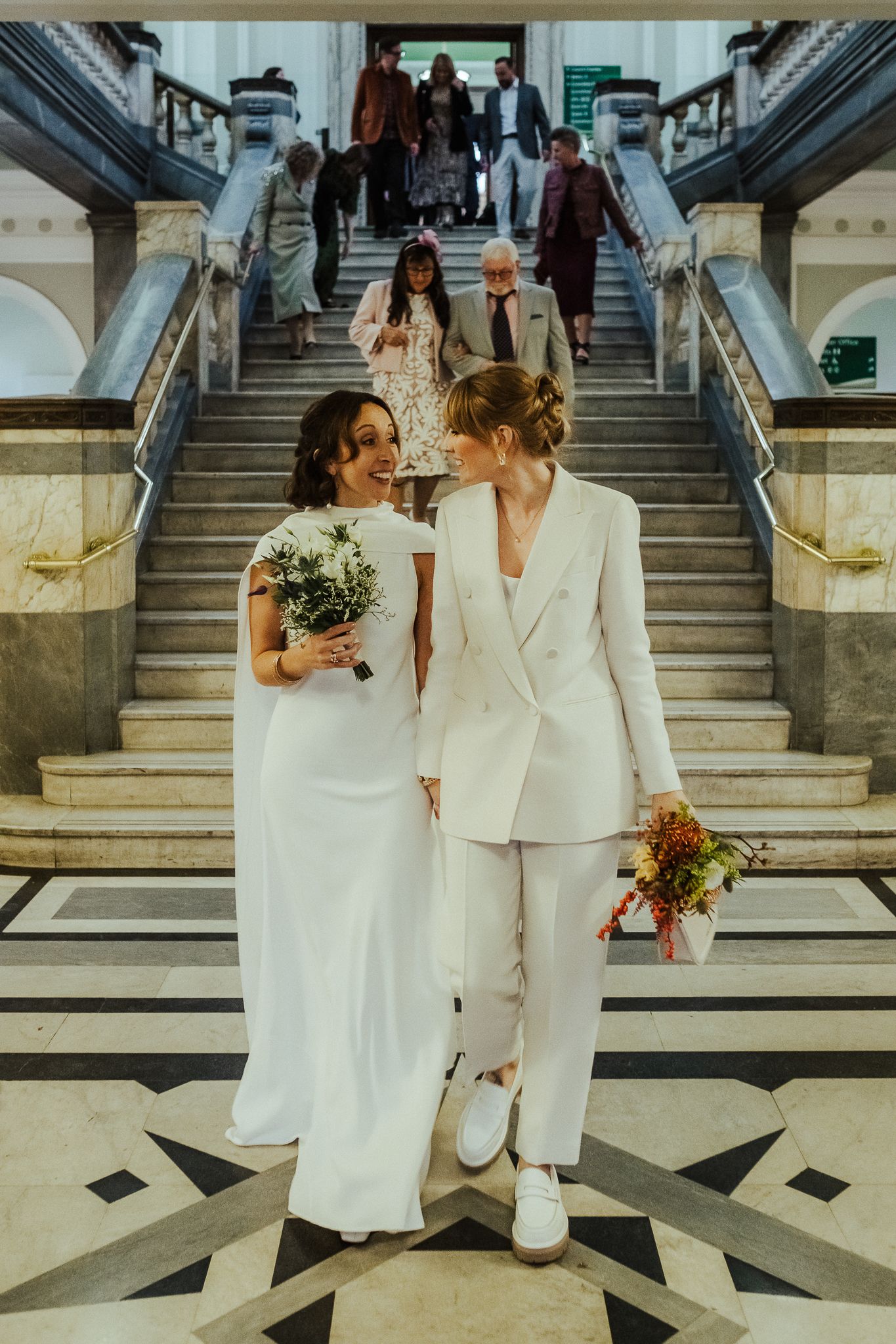 Two people in white wedding attire descend an elegant marble staircase while holding bouquets.