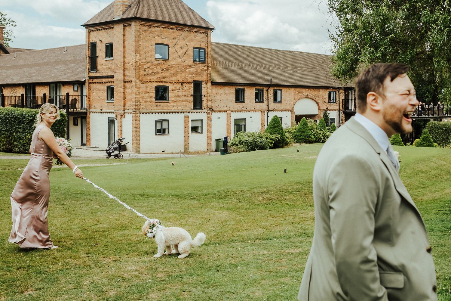 A scene outside a brick building with someone walking a small white dog on the lawn.