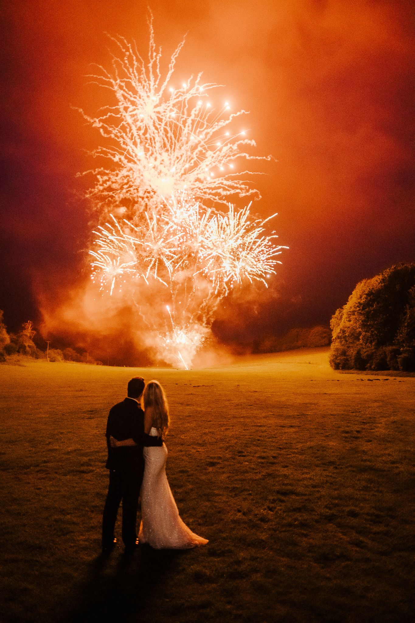 Spectacular fireworks display illuminates a couple against an orange night sky.
