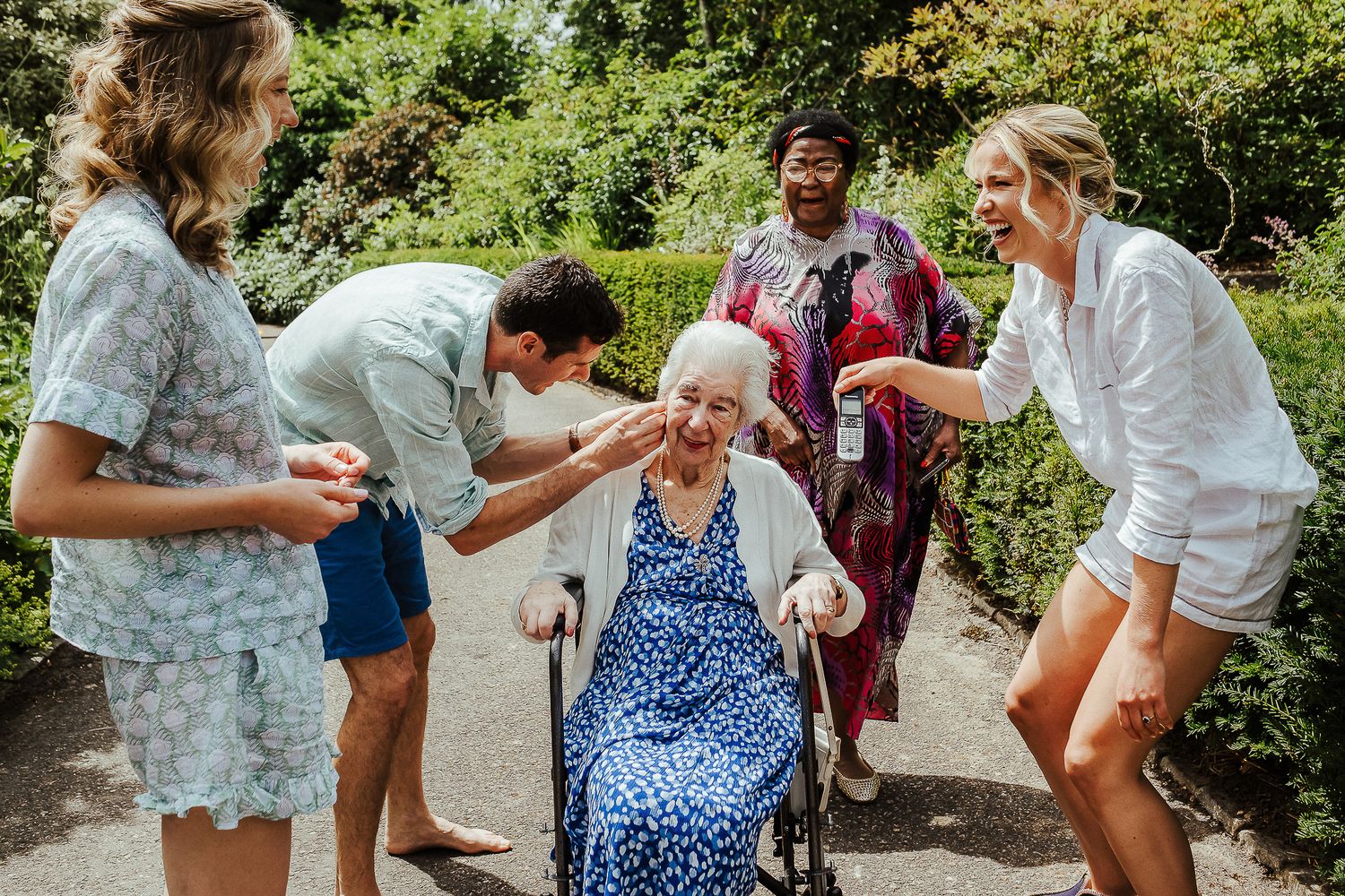 Medical staff and caregivers assist an elderly woman in a blue polka dot dress in an outdoor garden setting.