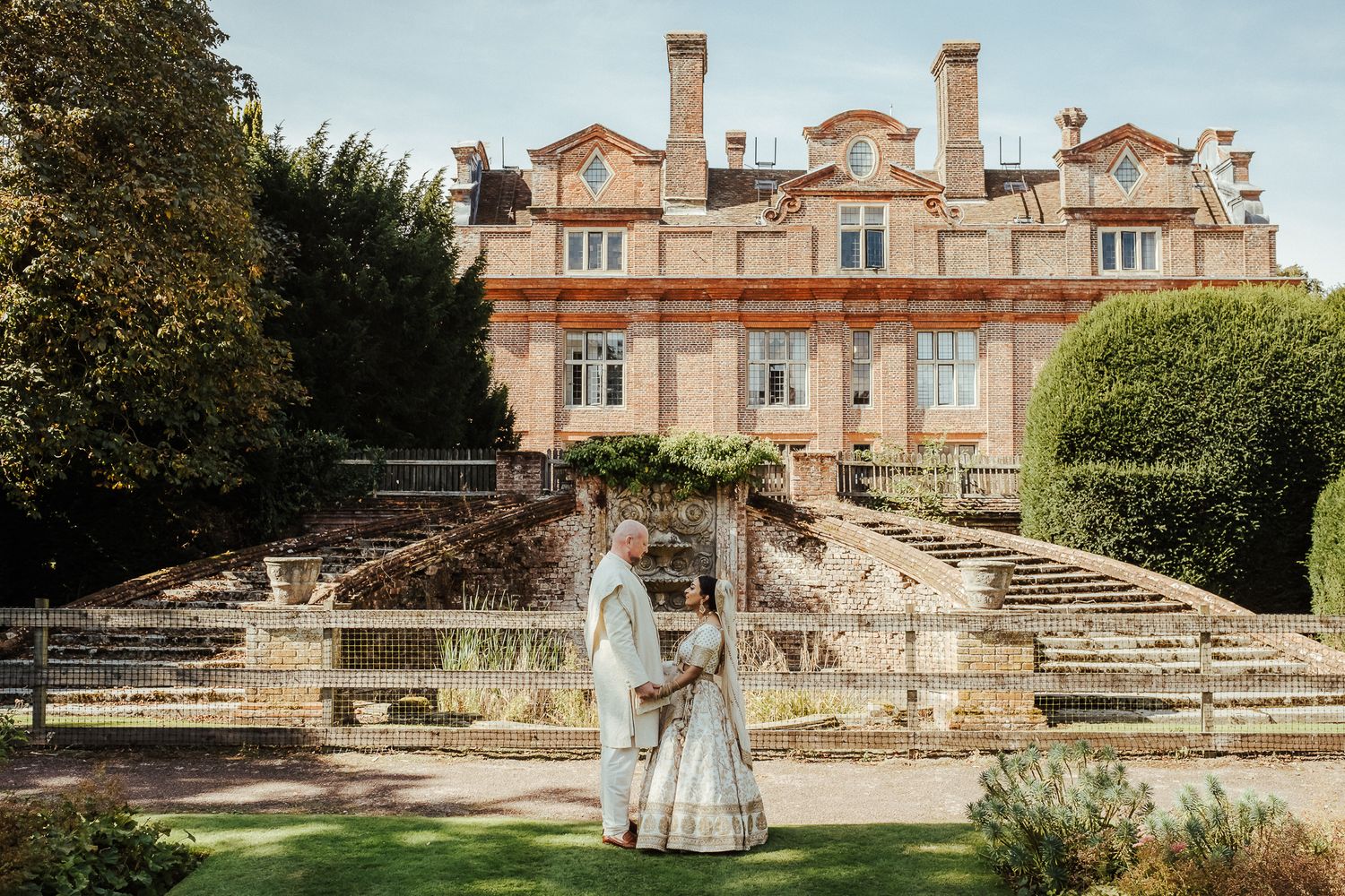 An elegant stone mansion with columned architecture stands behind manicured gardens with decorative fountains and staircases.
