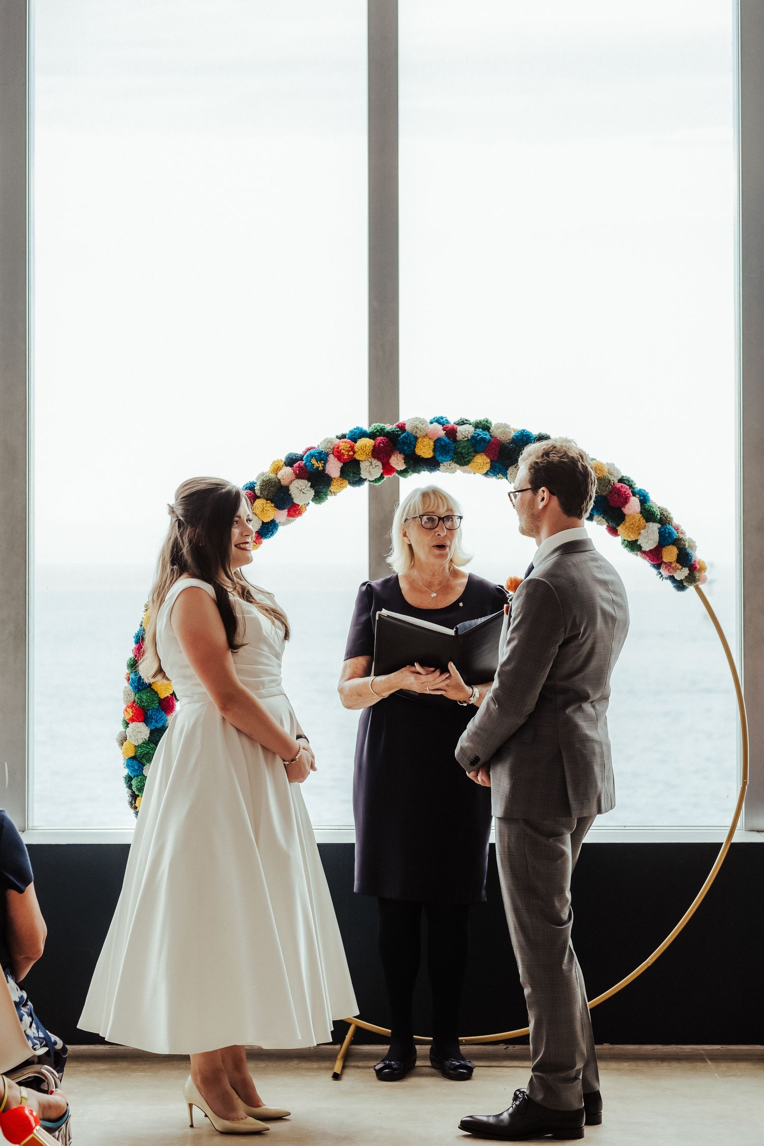 Couple exchanges vows under a colorful balloon arch during intimate indoor ceremony with ocean view.