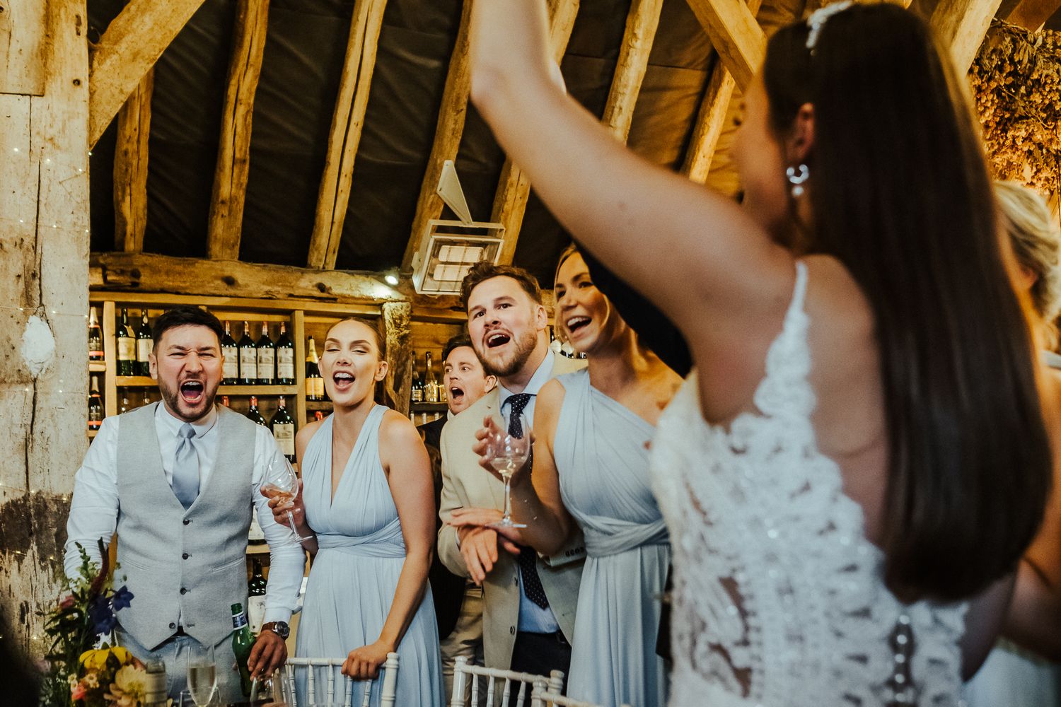 Wedding party members in gray and light colored attire dance and celebrate on a rustic wooden dance floor.
