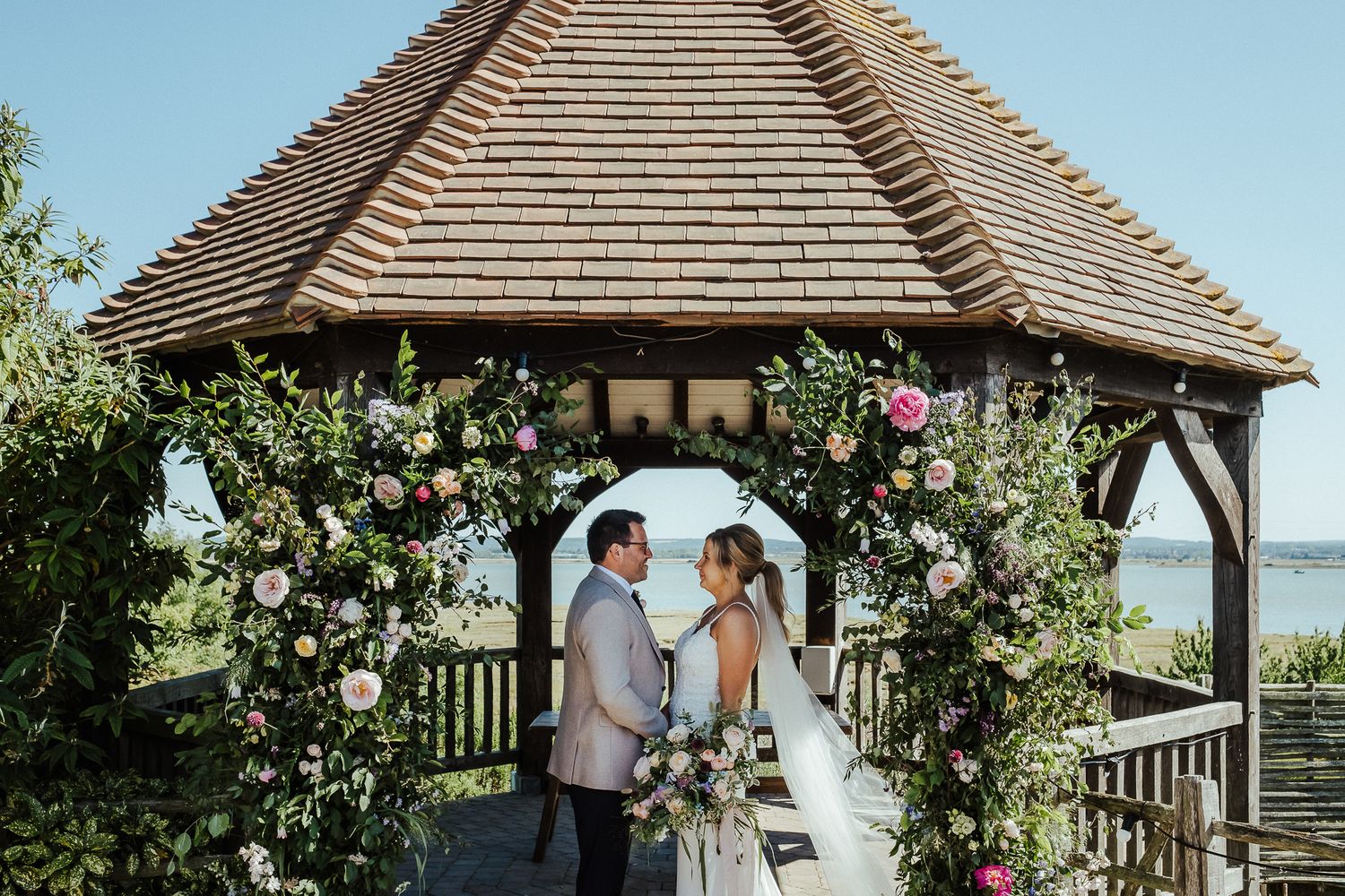 Couple stands in a rustic gazebo decorated with climbing roses and floral arrangements for their ceremony.