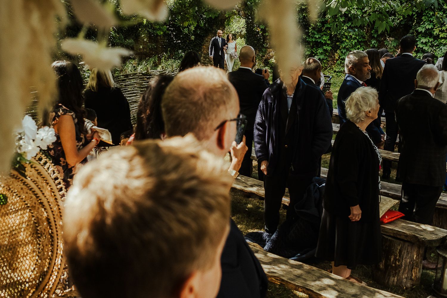Guests at an outdoor wedding ceremony viewed from behind with natural lighting.