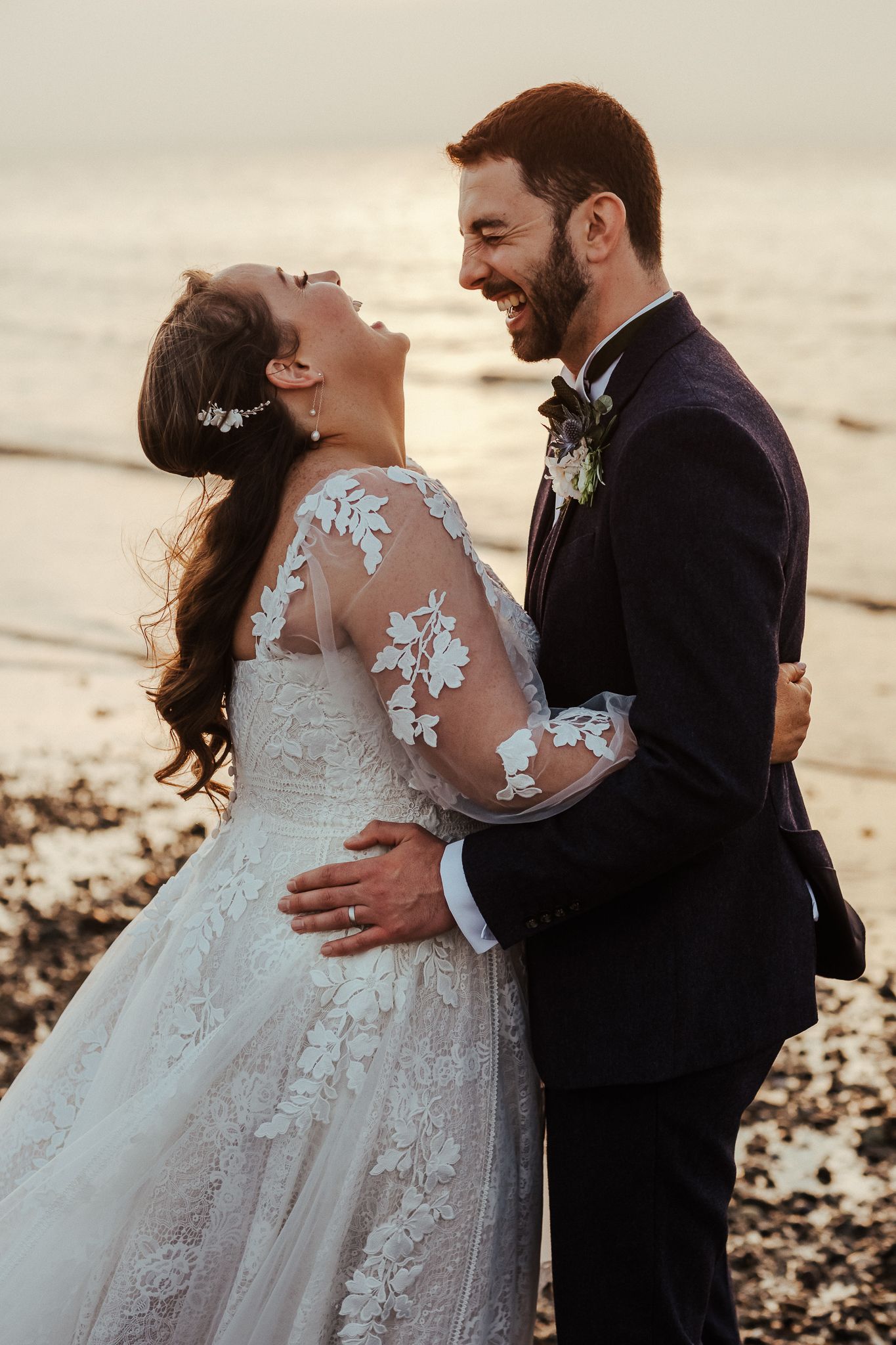 Couple shares intimate moment at sunset on rocky beach in wedding attire.