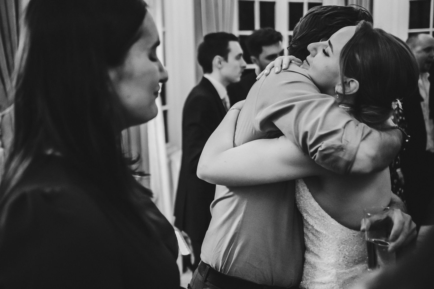 A black and white candid shot of guests embracing at a wedding reception indoors.