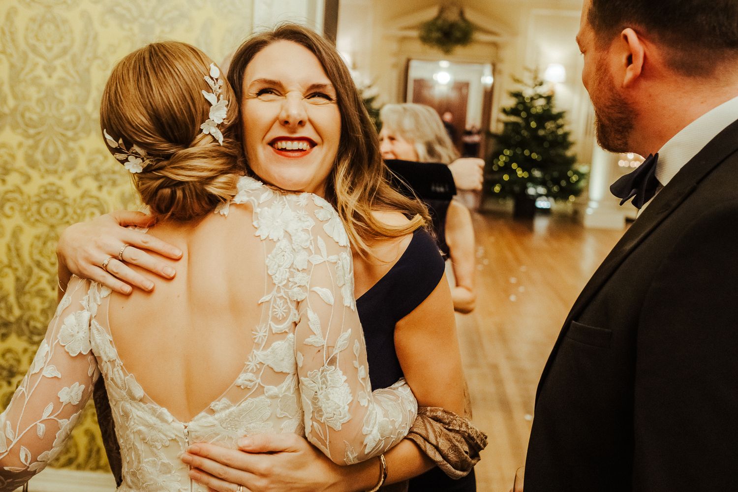 Emotional reception moment as guests share warm embraces in golden-lit room with Christmas tree.
