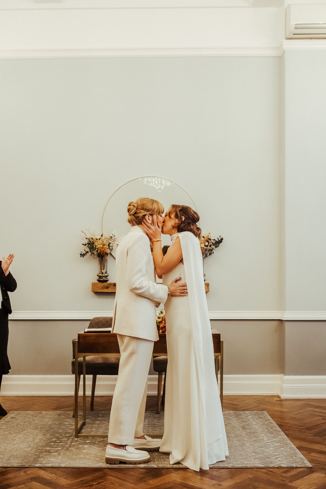 Couple in white suits sharing their first kiss under an archway during wedding ceremony.