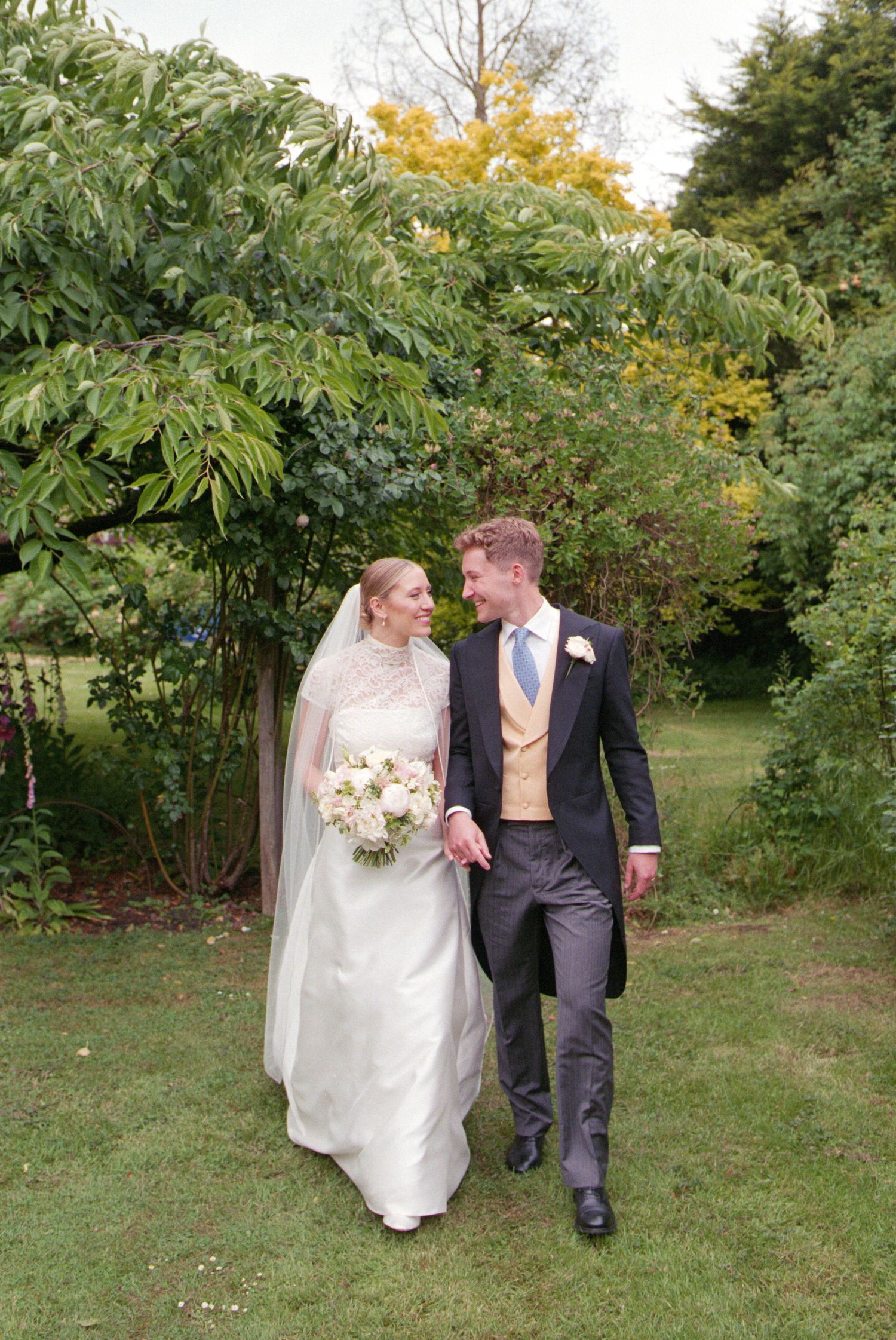 A couple walks together through a lush garden setting on their wedding day in autumn.