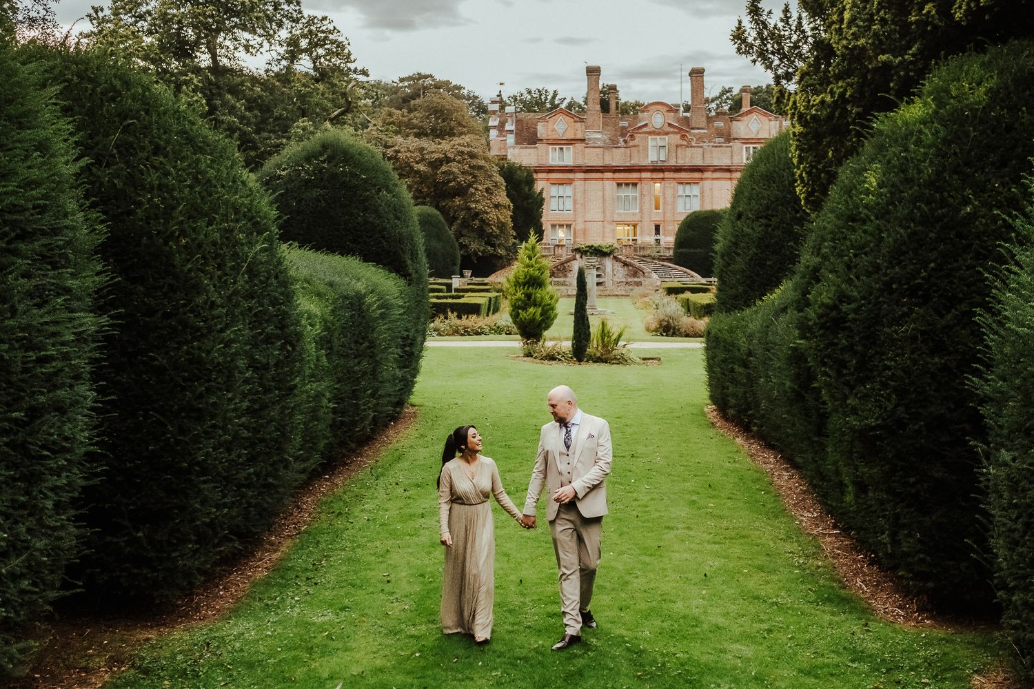 A couple walks down a manicured garden path lined with tall hedges leading to a stately brick manor house.