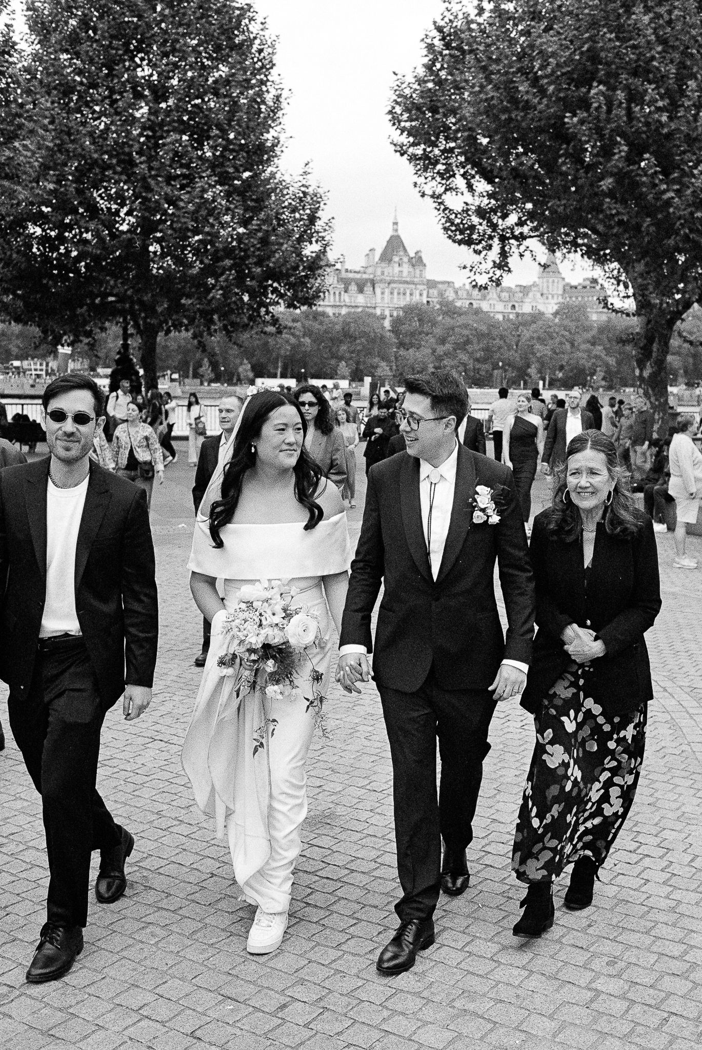 A wedding party walks along a brick path with large trees and a castle-like building visible in the background.