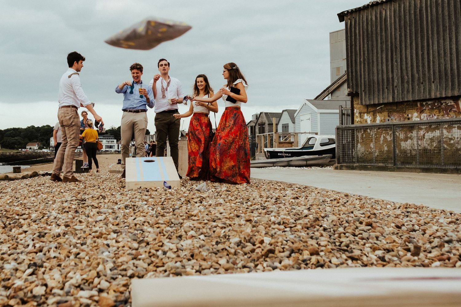 Wedding guests gather on a pebble beach enjoying drinks and conversation near a rustic wooden building.