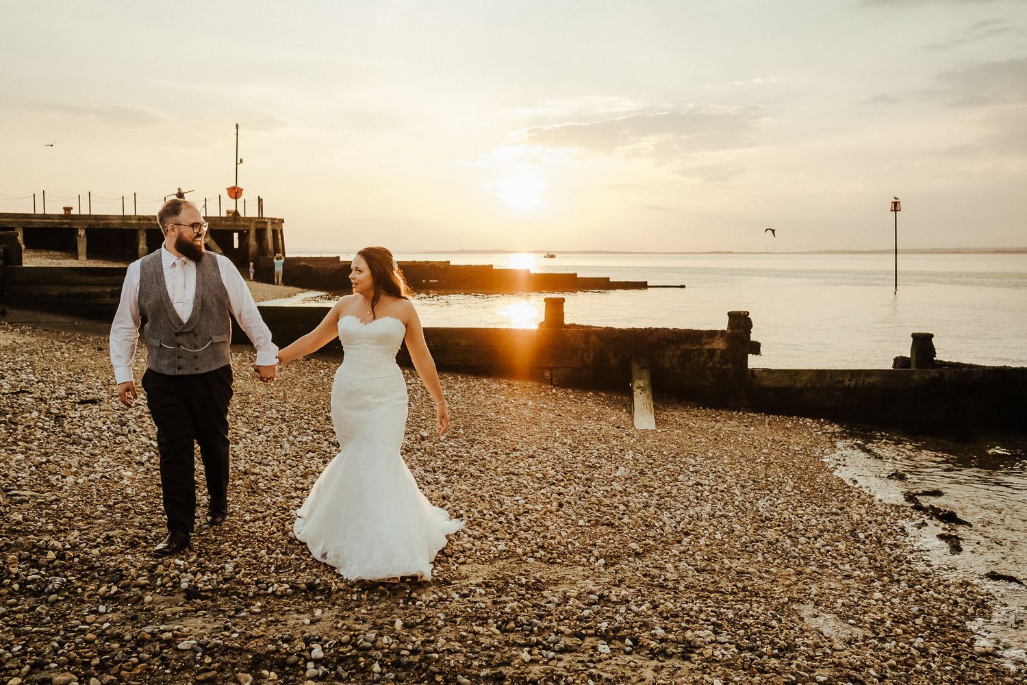 A couple walking along a beach at sunset with groynes and pier in the background.