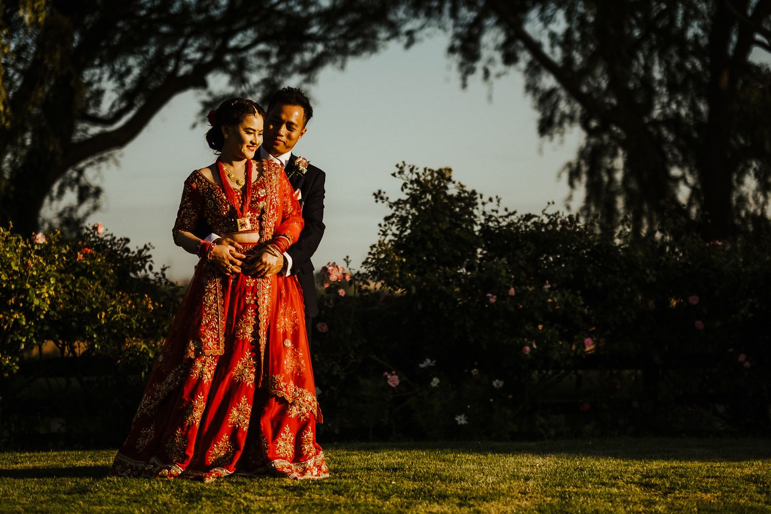 Couple in red traditional wedding attire poses during golden hour in a garden setting.