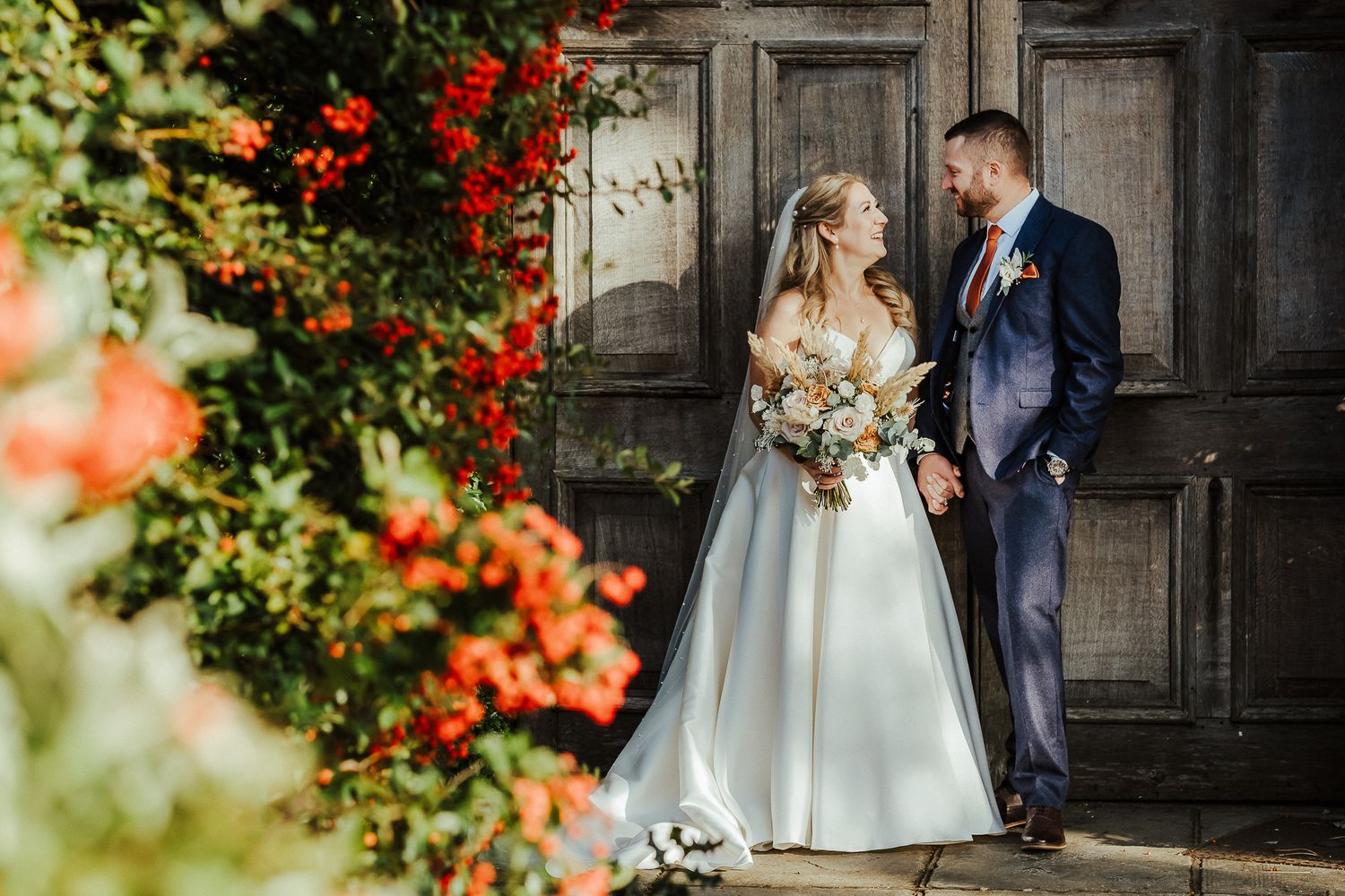 Elegant wedding couple shares intimate moment by rustic door with blooming red flowers.
