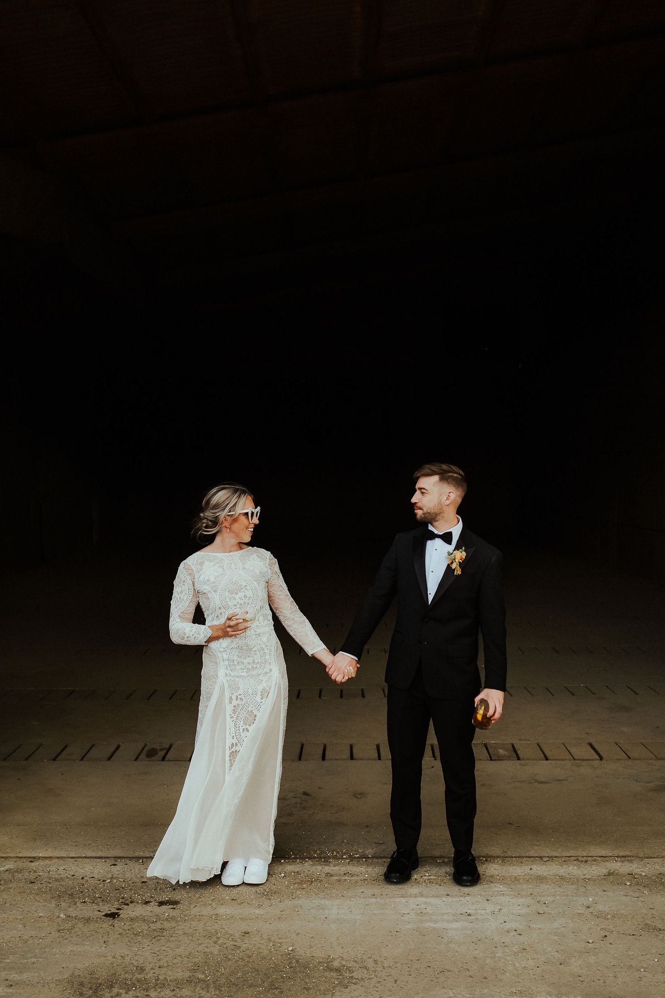 Wedding couple stands together in elegant attire against dark background.