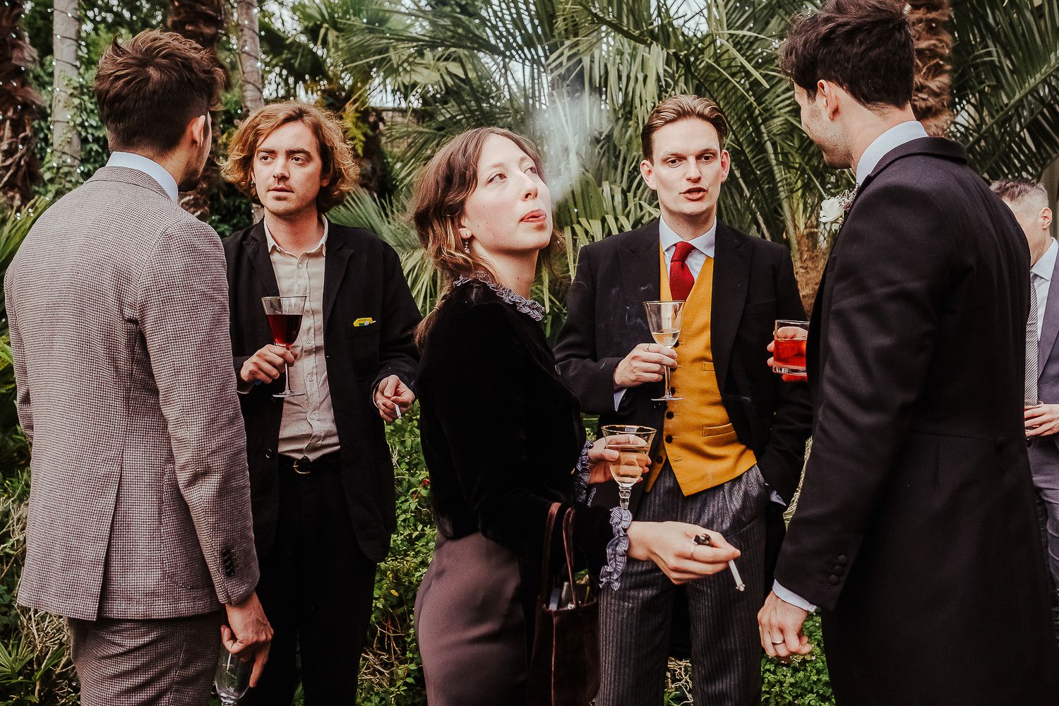 Group of elegantly dressed guests enjoying conversation at cocktail reception in conservatory setting.