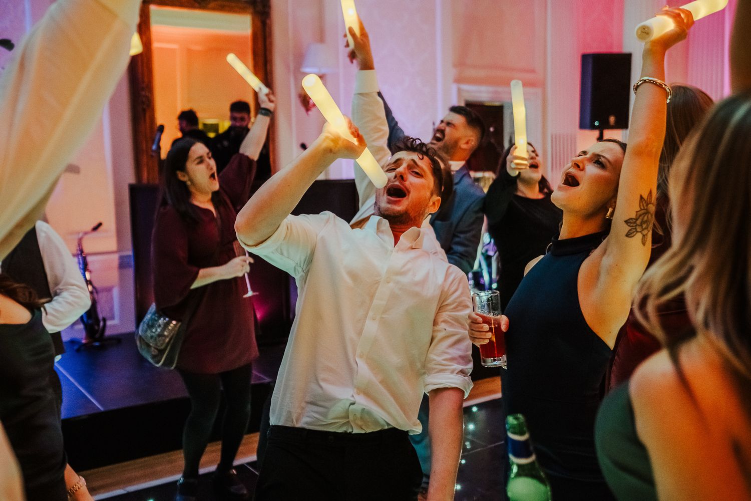 Wedding guests dancing energetically with raised arms under pink mood lighting at an indoor reception.