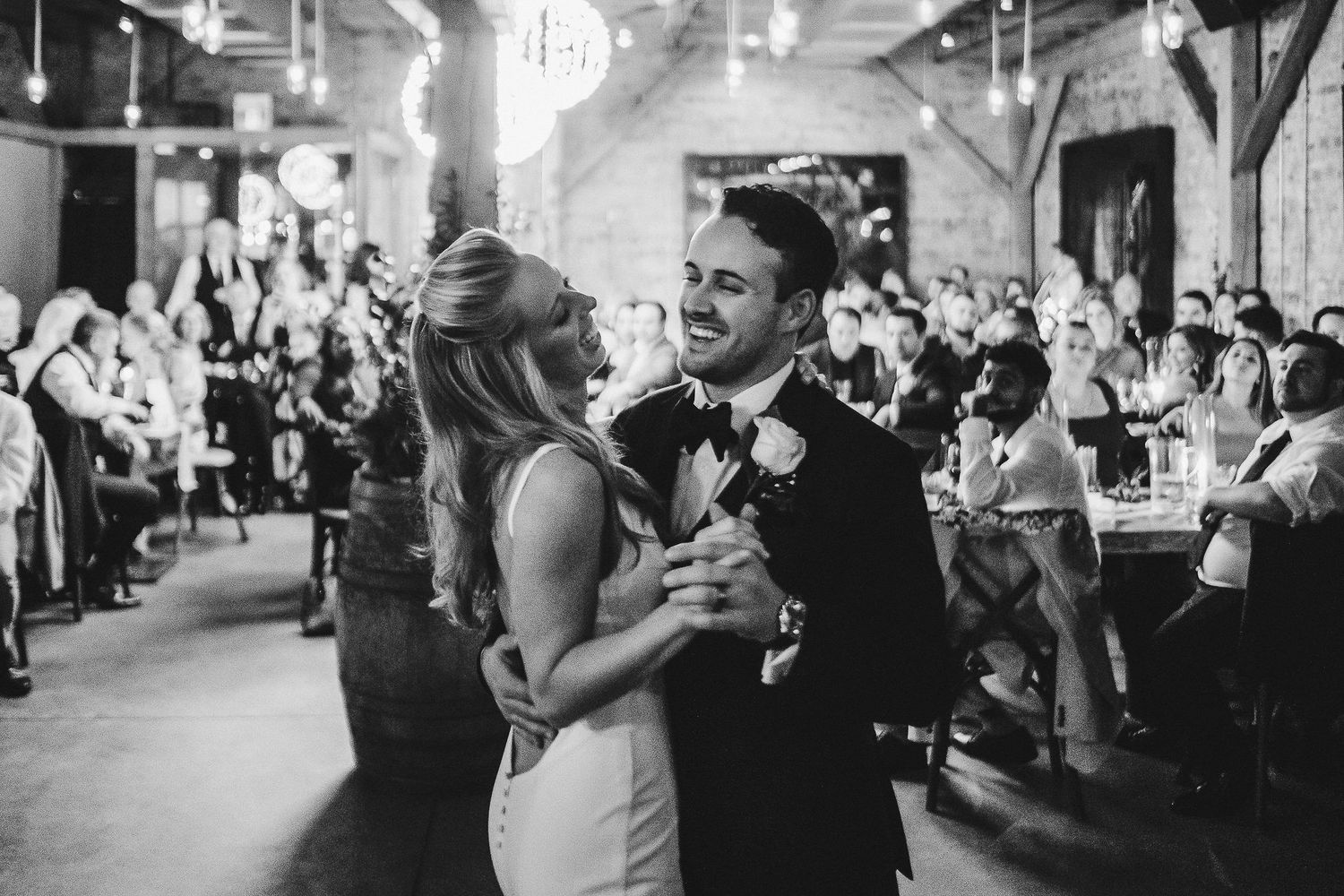 A couple dancing together in a rustic venue with guests watching in the background.
