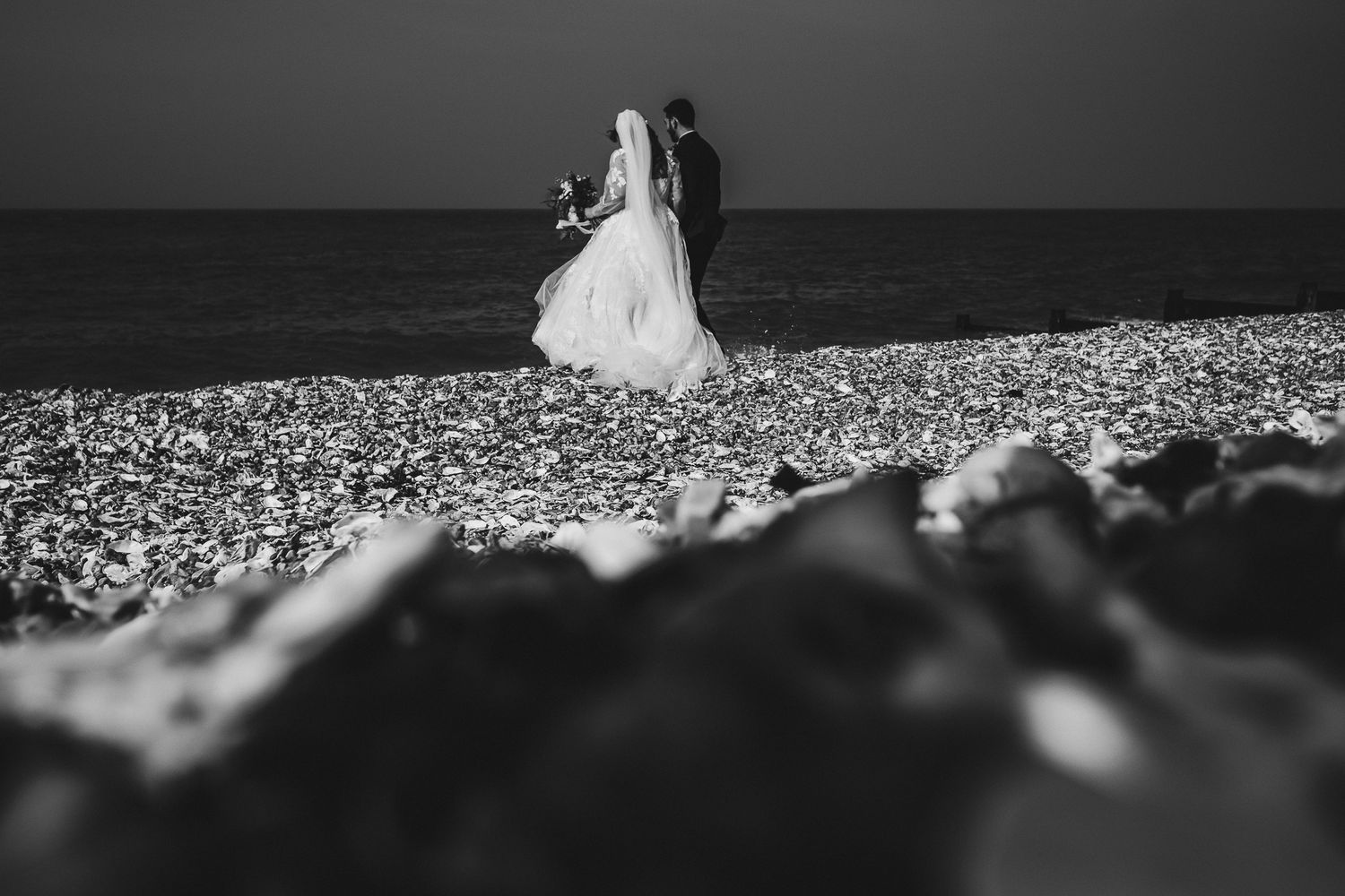Black and white silhouette of newlyweds standing on a rocky beach at sunset.