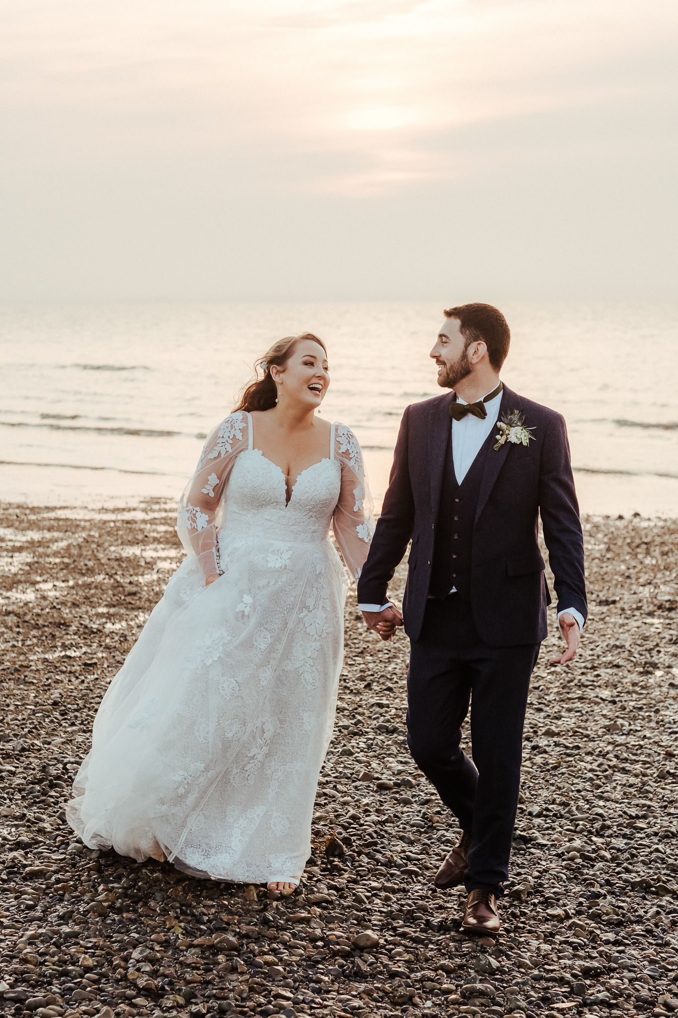 Happy couple walks along pebbly beach at sunset in elegant wedding attire.