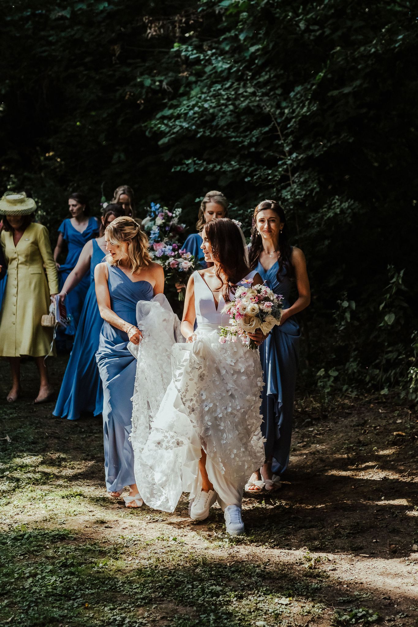 Bridesmaids in blue dresses help carry a wedding dress train along a forest path.