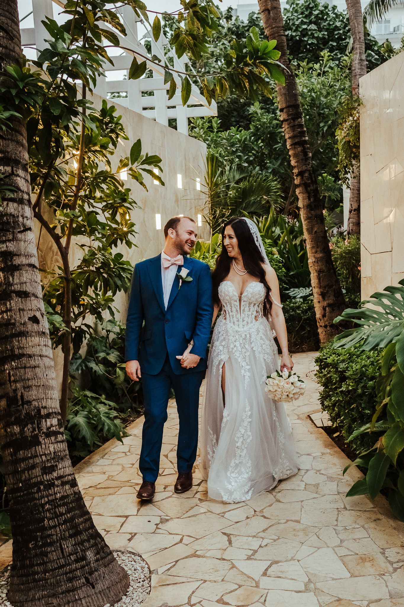 Bride in lace gown and groom in blue suit stroll through tropical garden pathway.