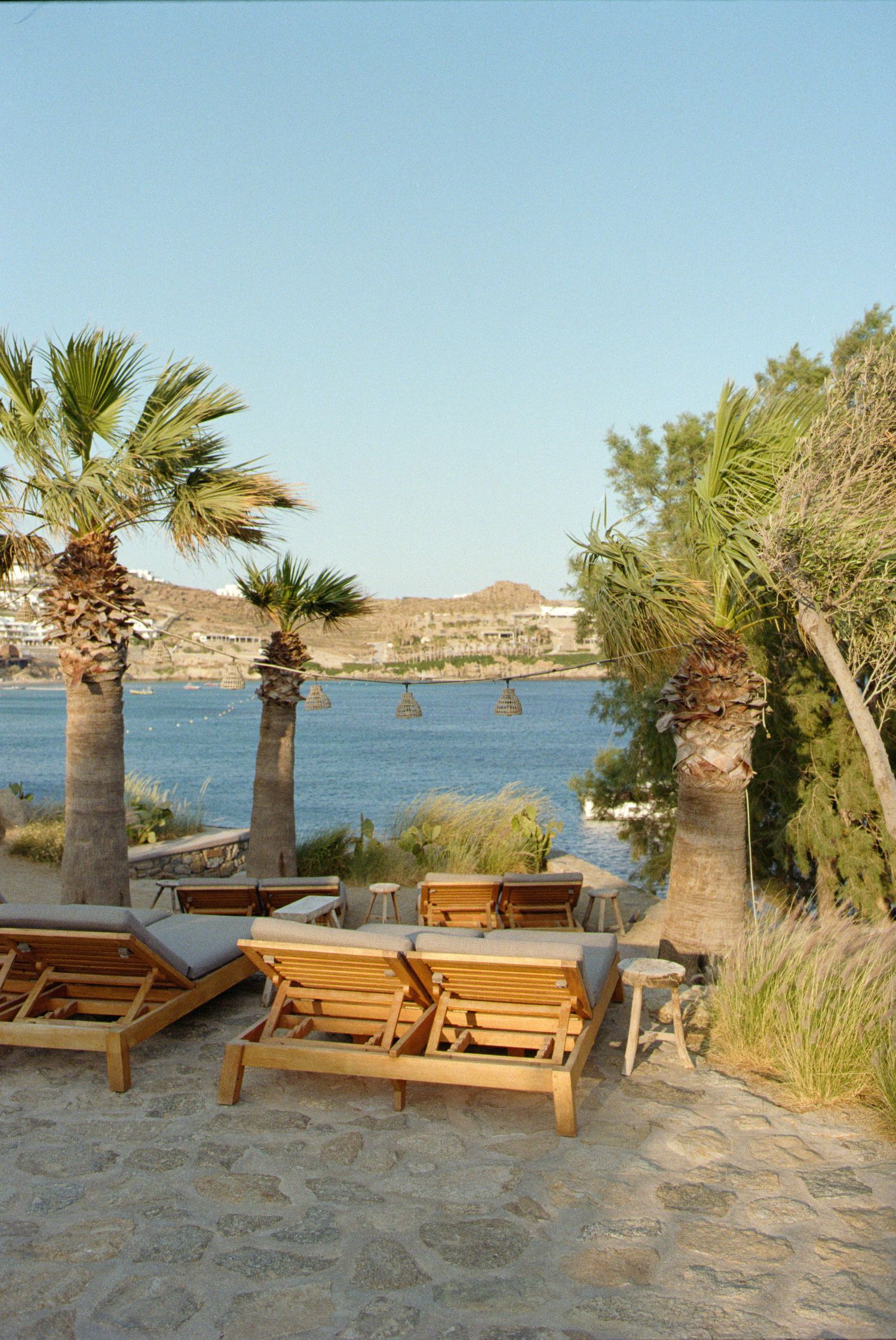 Wooden lounge chairs and palm trees overlook a Mediterranean bay with rocky coastline and clear blue waters.